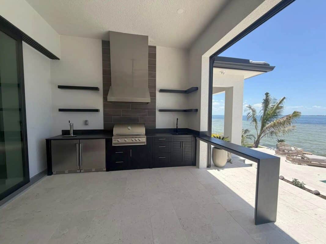 Waterfront outdoor kitchen with dark cabinetry, built-in grill and covered patio overlooking the bay